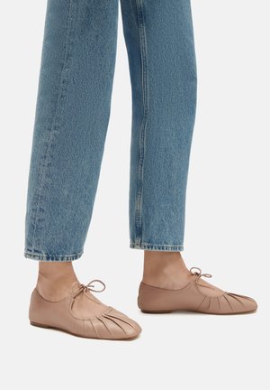 Világos barna bőr cipők redőzött dizájnnal, kis masni részlettel és lapos talppal, amelyeket közepes derekú kék farmernadrággal viselnek.