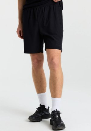 Mand iført sorte atletikshorts, hvide ankelsokker og sorte sneakers, står på en hvid baggrund.