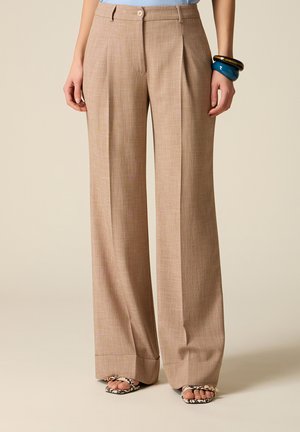 Pantaloni beige a gamba larga con pieghe frontali, orli risvoltati, indossati con sandali aperti con stampa serpente e braccialetti spessi blu e oro.