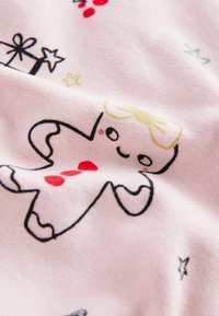Mīksts rozā tekstils ar piparkūku cilvēciņa ilustrāciju, kuram ir dzeltena loksne, sarkani pogas un dažādi dāvanām un zvaigznēm raksti tumšā krāsā.