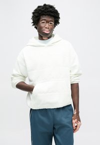 Sweat à capuche en polaire blanche avec une grande poche avant, présentant un logo embossé. Associé à un pantalon turquoise, pour un look décontracté et sportif.