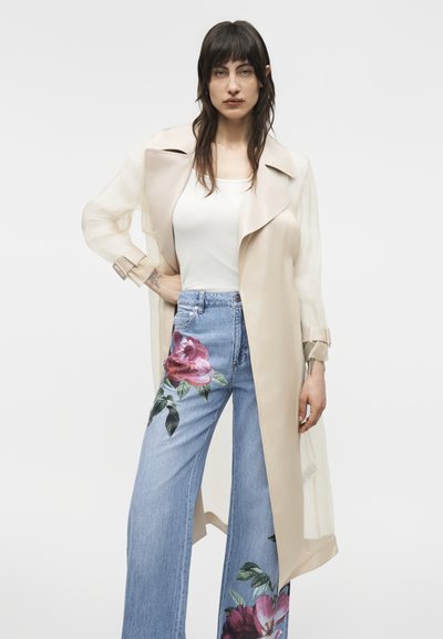 Žena v priehľadnom béžovom trenčkotu, bielom topánku a svetlomodrých rifle s vysokým pásom a veľkými ružovými kvetinovými vzormi, stojaca pred jednoduchým pozadím.