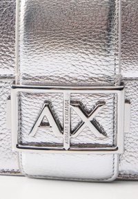 Srebrna teksturirana torbica z rektangularno sponko, ki nosi logotip in blagovno znamko "AX", ki prikazuje eleganten in sodoben dizajn.