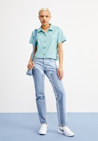 O cămașă cu mânecă scurtă, de culoare turquoise, cu un model decorativ, asociată cu blugi deschiși la culoare și adidași albi cu accente verzi.