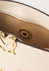 Τσάντα από δέρμα με υφή κρέμας, με κούμπωμα σε σχήμα χρυσής καρδιάς και εσωτερική στρογγυλή χρυσή ετικέτα που αναγράφει "LOVE MOSCHINO" με σύμβολα καρδιάς.