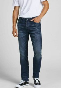 Mörkblå denimjeans med figursydd design, ljus slitage framtill, femficksstil och metallknäppning. Matchas med svarta sneakers.