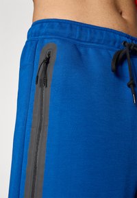 As calças atléticas azuis apresentam uma cintura com cordão, bolsos laterais com zíper e detalhes cinzentos, além de um tecido liso e texturizado.