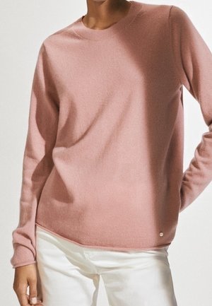 Persoon draagt een zachtroze trui met lange mouwen en witte broek, poseert met één hand op de heup tegen een effen achtergrond.