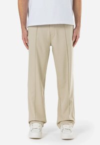 Pantalon beige en tissu lisse avec une coupe droite. Présente des détails de couture visibles et une coupe décontractée, associé à des baskets blanches.