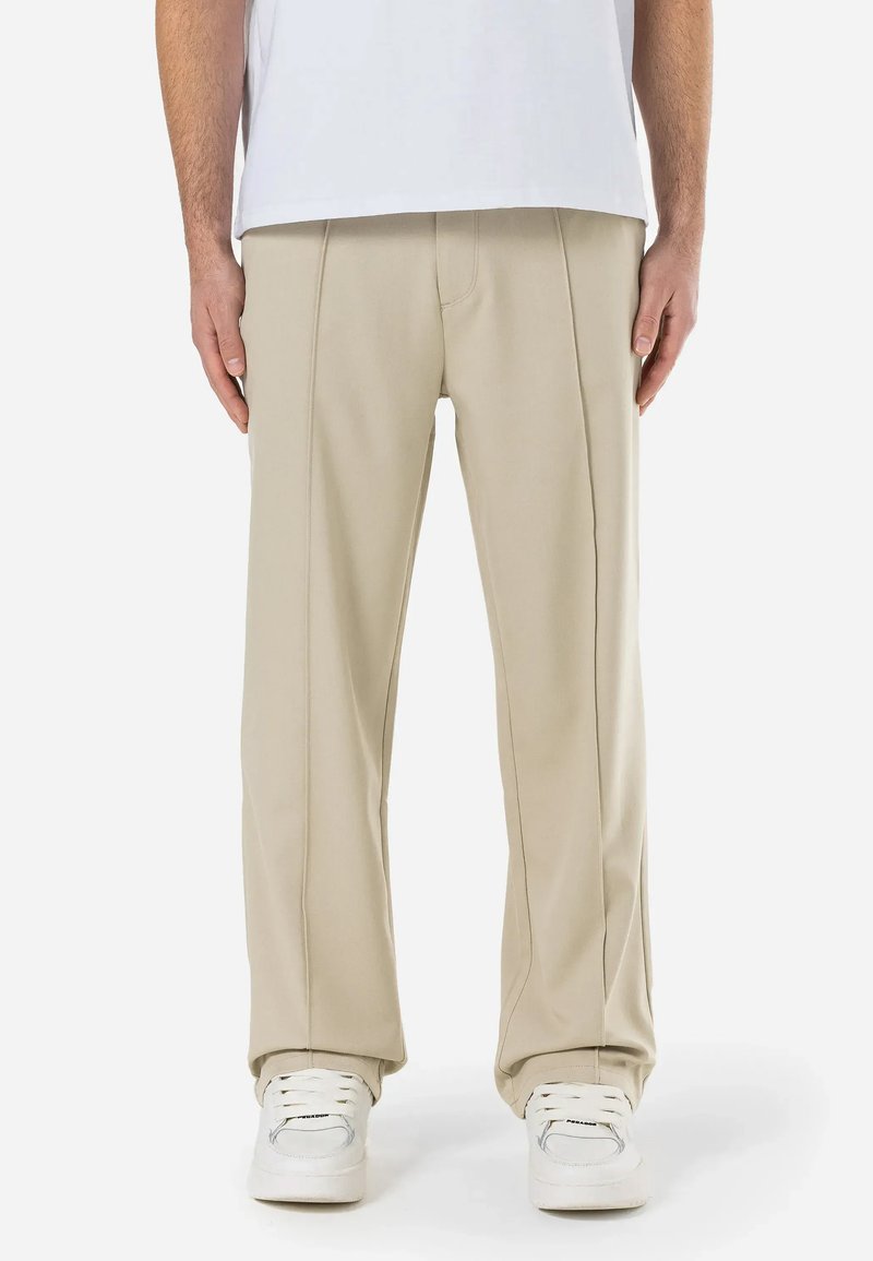 Pantalon beige en tissu lisse avec une coupe droite. Présente des détails de couture visibles et une coupe décontractée, associé à des baskets blanches.