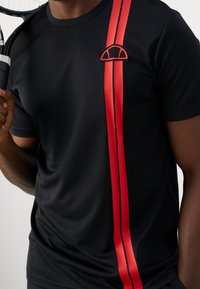 Camiseta atlética negra con dos rayas rojas verticales y un pequeño logo en el pecho. Tejido suave con un diseño ajustado.