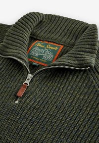 Pull en laine vert avec col renforcé et fermeture éclair, portant une étiquette "Next Supply" en orange, et une fermeture éclair en argent avec accent en cuir.
