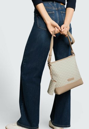 Person wearing dark blue jeans and white shoes holds beige patterned handbag behind back against plain background.