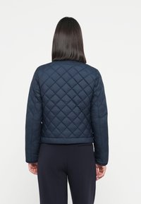 Marinefarget quilted puffer-jakke med ribbestrikket kant og lange ermer. Har et diamantmønster og glatt tekstur. Vist bakfra.