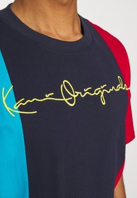 Námořnické tričko s červenými a tyrkysovými rukávy, s žlutým kurzivním vyšitým nápisem "Kani Origindal." Krátký rukáv, bavlněný materiál.