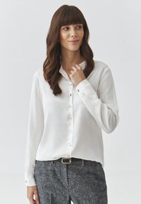 Biała koszula zapinana na guziki z kołnierzykiem, długimi rękawami oraz zielonymi akcentami przy guzikach. Zestawiona z szarymi spodniami o fakturze. Czysty i elegancki design.