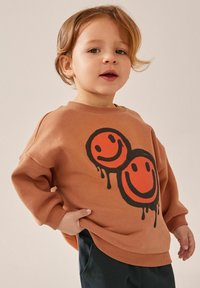 Brązowy sweatshirt z dwiema grafikami uśmiechniętych twarzy w kolorze pomarańczowym i czarnym. Miękka tkanina, okrągły dekolt, ściągacze przy mankietach oraz luźny krój.