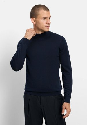 Uomo con capelli corti che indossa un maglione dolcevita blu scuro e pantaloni neri, che sistema il colletto mentre guarda a destra.