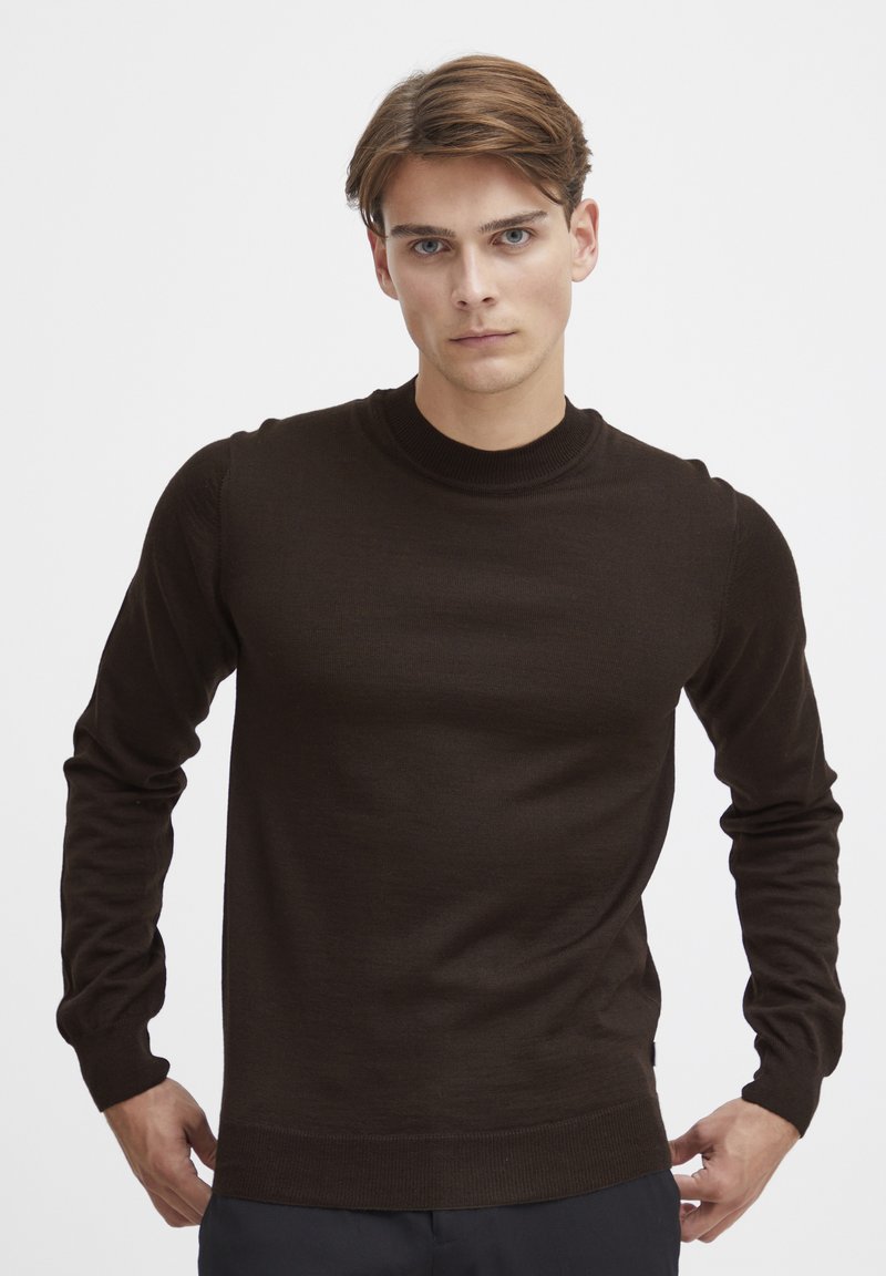 Pullover marrone lavorato a maglia con maniche lunghe e colletto alto. Texture liscia con una silhouette aderente e polsini e orlo a coste.