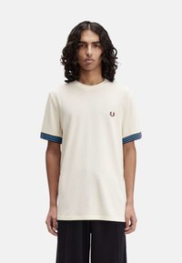 Camiseta color crema de manga corta, con detalles en rayas azul y negra en los puños y un pequeño emblema bordado en el pecho.