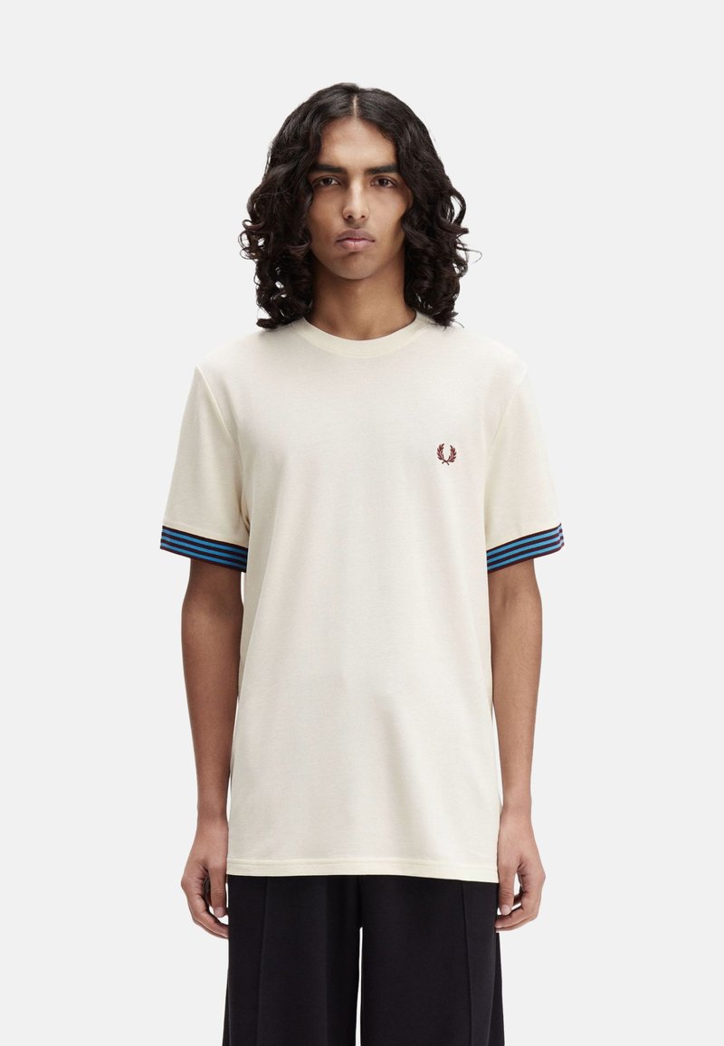 Camiseta color crema de manga corta, con detalles en rayas azul y negra en los puños y un pequeño emblema bordado en el pecho.