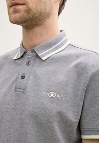 Graues Poloshirt mit strukturiertem Stoff, dreiknöpfiger Knopfleiste und gestreiften Kragenakzenten in Weiß und Grün. Kleines Logo mit "1962" gestickt.