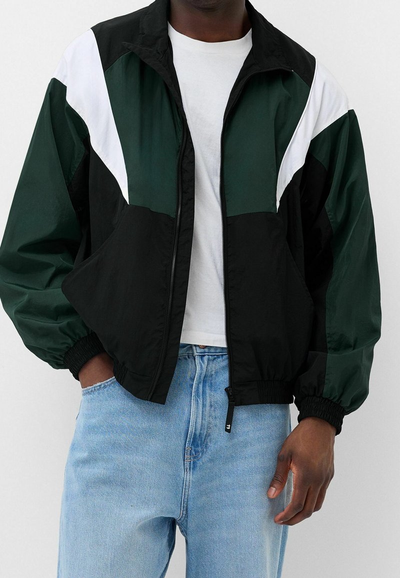 Färgblockerad jacka med svarta, mörkgröna och vita paneler, tillverkad av lättviktsmaterial med dragkedja fram och elastiska ärmslut.