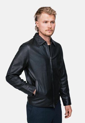 Jeune homme aux cheveux clairs portant une veste en cuir noire et un pantalon sombre, debout avec une main dans la poche sur un fond uni.