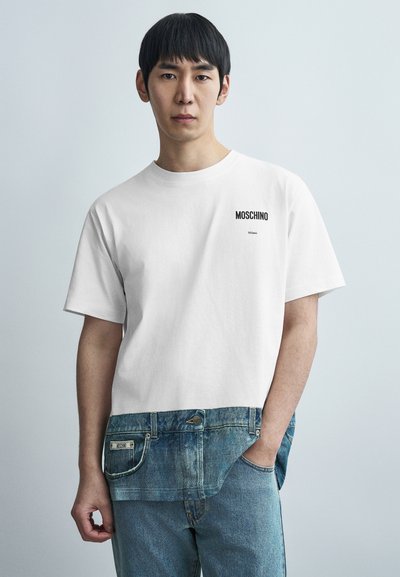 T-shirt en coton blanc avec un panneau en denim à l'ourlet, portant les inscriptions « MOSCHINO » et « Milano » en texte noir sur la poitrine.