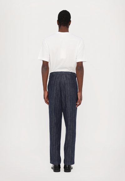 Donkerblauwe pantalons met een rechte pasvorm, gedragen met een effen witte korte mouwen shirt. Het perspectief is van achteren, waardoor het op maat gemaakte ontwerp van de broek opvalt.