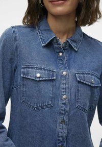 Blue denim shirt with a collared neck, button-up front, and two chest pockets. Features metal buttons and a textured fabric finish.