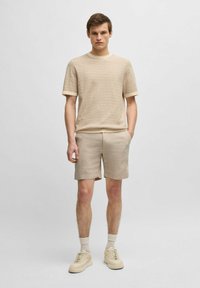 Maglione beige a maniche corte con una texture, abbinato a pantaloni corti in lino beige e sneakers crema. Calzini chiari completano l'outfit.