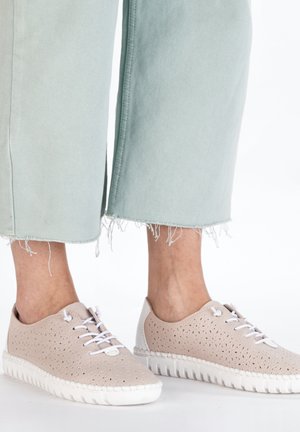 Baskets perforées beiges avec semelles blanches, présentant six œillets et des accents de talon blancs contrastants. Stylisées avec un jean court vert clair.