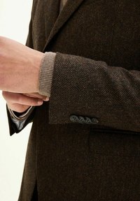 Blazer a spina di pesce marrone scuro con bottoni neri, con un polsino beige a coste che spunta sotto la manica. Tessuto di lana testurizzato.