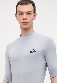 Világosszürke, rövid ujjú rash guard, fekete logóval a mellkason és minimális varrási részletekkel. Rugalmas anyagból készült.