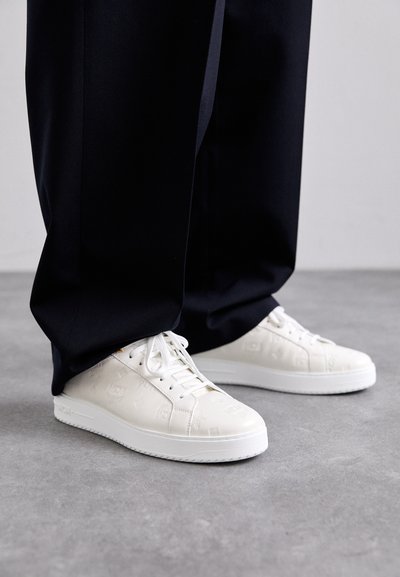 Zapatillas blancas con patrones de logotipos en relieve, suelas de goma y cordones blancos, combinadas con pantalones oscuros y holgados.