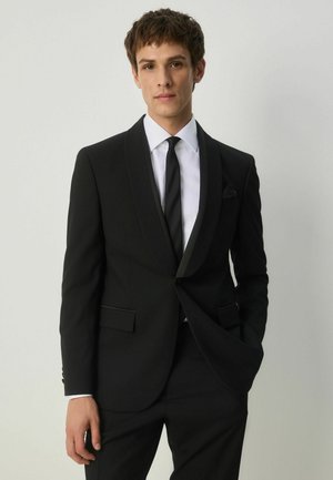 Jeune homme portant un smoking noir avec des revers en satin, une chemise blanche et une cravate noire, se tenant devant un fond clair uni.