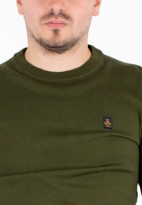 Man met een korte baard die een olijfgroen gebreid trui draagt met een klein zwart en oranje logo op de borst.