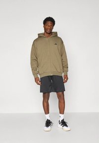 Khaki glidelåshoodie med hette, med svart logo og ribbet kant, kombinert med svarte shorts og hvite treningssko.