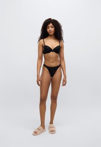Σετ μαύρα εσώρουχα με κυματιστές άκρες, σε συνδυασμό με μπεζ χνουδωτά παντόφλες. Το μοντέλο στέκεται μπροστά σε ανοιχτό γκρι φόντο.