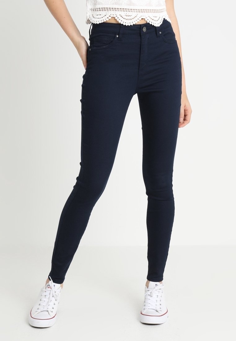 Femme portant un jean skinny bleu foncé, un crop top en dentelle blanche et des baskets blanches, se tenant avec une main dans la poche devant un fond blanc.
