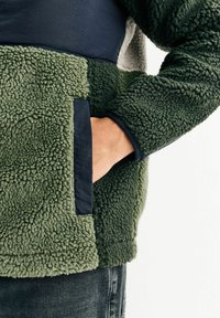Fleece-pullover med struktureret grøn krop, marineblå detaljer og en påsyet lomme. Har en krave-halvkrave og en blød, plysset finish.
