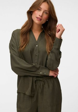 Jeune femme aux longs cheveux bruns portant une blouse boutonnée vert olive et un pantalon à cordon de serrage assorti sur fond blanc.