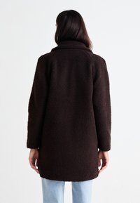 Manteau en fausse fourrure texturée marron avec un large col, des manches longues et une coupe ample, s'étendant jusqu'à mi-cuisse, porté sur un jean en denim clair.