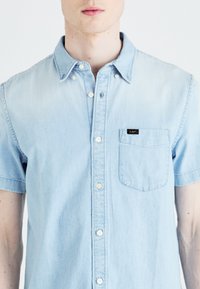 Chemise en denim à manches courtes de couleur bleu clair, avec une poche à rabat et une petite étiquette noire sur la poitrine gauche.