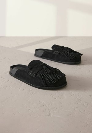 Czarne zamszowe buty wsuwane na płaskiej podeszwie z okrągłym noskiem, ozdobione dekoracyjnym frędzlem i detalami ściegów na cholewce. Gładka, płaska podeszwa.