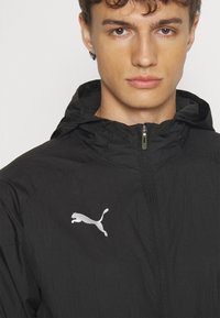 Jonge man in een zwart gewatteerd jack met capuchon en een zilveren Puma-logo op de linkerborst, die recht vooruit kijkt tegen een effen achtergrond.