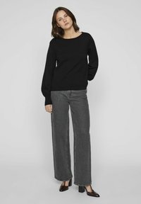 Un pull noir avec un col large, des manches longues et des poignets côtelés, associé à un jean gris à taille haute et jambes droites, avec des chaussures à talons carrés.