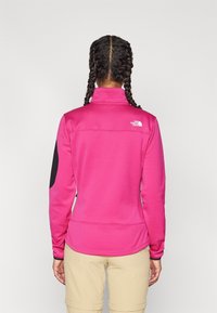 Fleece pulovr v jasně růžové barvě s polovičním zipem, černými ramenními akcenty a malým bílým logem na horní části zad. Hladká textura.