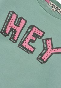 Grüner Hoodie mit auffälligem "HEY"-Text in Pink, versehen mit gestickten Pailletten und einer grauen Umrandung. Der Stoff ist weich und strukturiert.